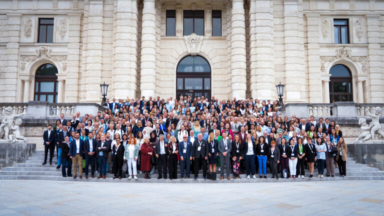 The picture of a big group of people standing on the stairs outside of The Hofburg, Vienna. They are wearing casual business clothes and they are all smiling at the camera.